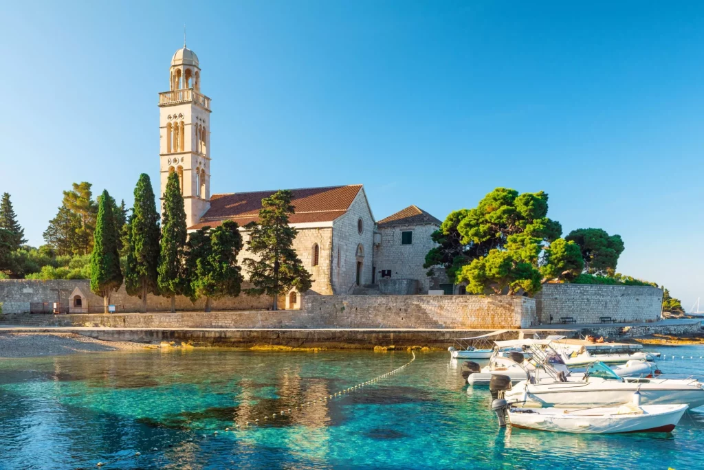 Türkisfarbenes Wasser der Adria-Bucht auf der Insel Hvar mit Franziskanerkloster und Booten in der Region Dalmatien, Kroatien bei Sonnenaufgang